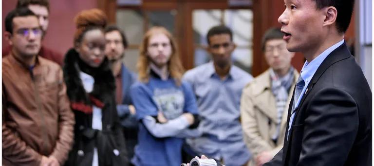 An east asian man in a suit is in focus on the right talking a group of people.